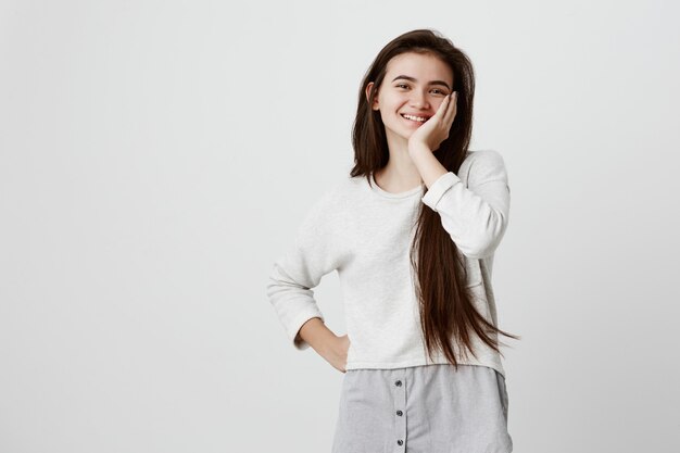 Retrato en la cintura de la modelo femenina morena sonriendo alegremente con dientes blancos, contento de recibir buenas noticias o presentes. La mujer emocionada expresa emotinos positivos, sostiene la mano en la mejilla con asombro.