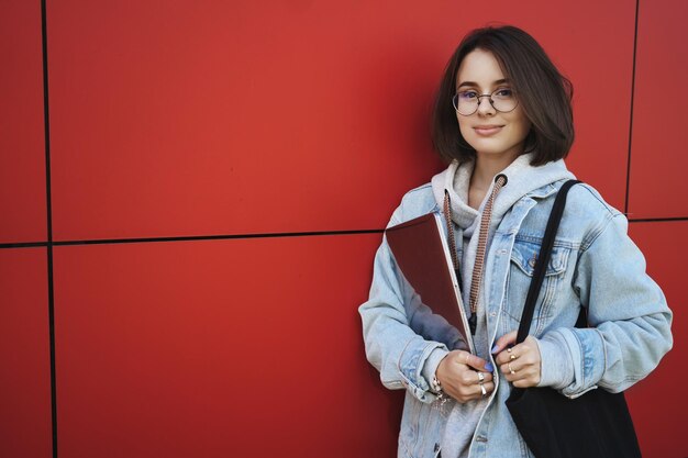 Retrato en la cintura de una mujer bastante caucásica segura de sí misma con gafas de chaqueta de mezclilla de pie sobre la pared del edificio rojo, sostenga una computadora portátil y una bolsa de mano, chatee rápidamente con un amigo al aire libre, cámara de sonrisa