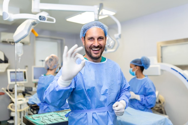 Retrato de un cirujano masculino en el quirófano mirando la cámara que muestra el gesto OK Doctor en exfoliantes y máscara médica en el quirófano de un hospital moderno