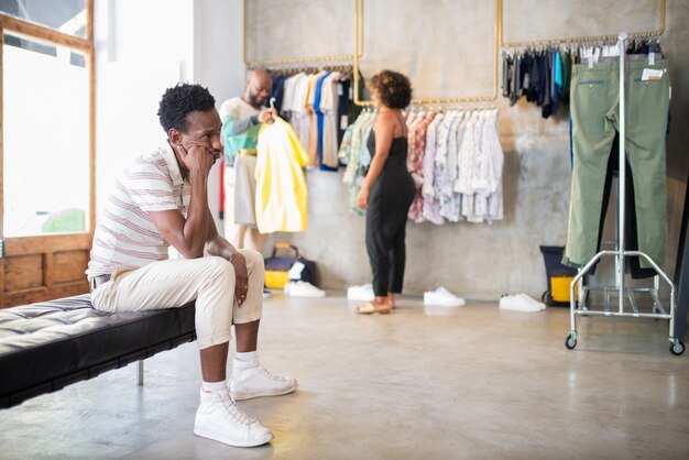 Retrato de clientes afroamericanos y vendedor en boutique. Mujer joven ayudando a un hombre barbudo a elegir una camisa mientras otro hombre sentado en el banco espera. Negocio boutique de ropa, concepto de compras