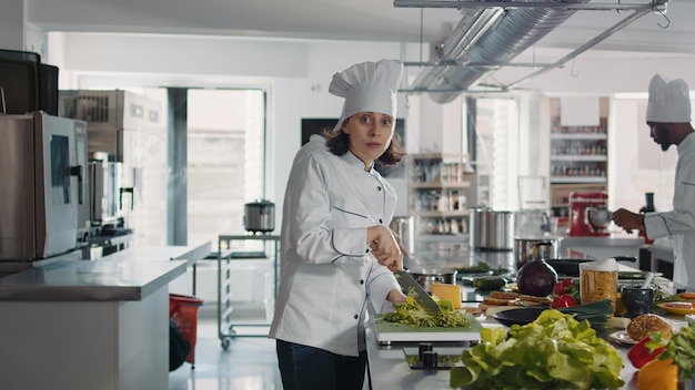 Retrato de una cocinera cortando apio verde en una cocina gourmet, usando una tabla de cortar y un cuchillo para cortar la ensalada de col. Mujer chef con uniforme preparando ingredientes para recetas de comida.