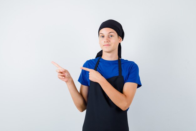 Retrato de cocinero adolescente masculino apuntando hacia el lado izquierdo en camiseta, delantal y mirando confiado vista frontal