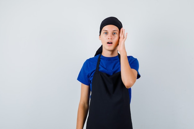 Retrato de cocinero adolescente masculino con la mano en la cabeza, abriendo la boca en la camiseta, delantal y mirando sorprendido vista frontal