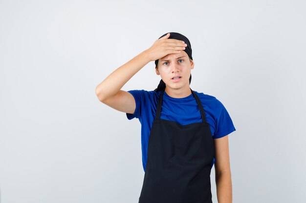Retrato de cocinero adolescente masculino manteniendo la mano en la frente en camiseta, delantal y mirando estresado vista frontal