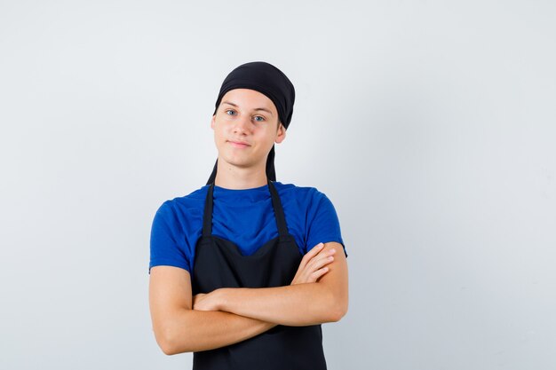 Retrato de cocinero adolescente masculino manteniendo las manos cruzadas en la camiseta, delantal y mirando confiado vista frontal