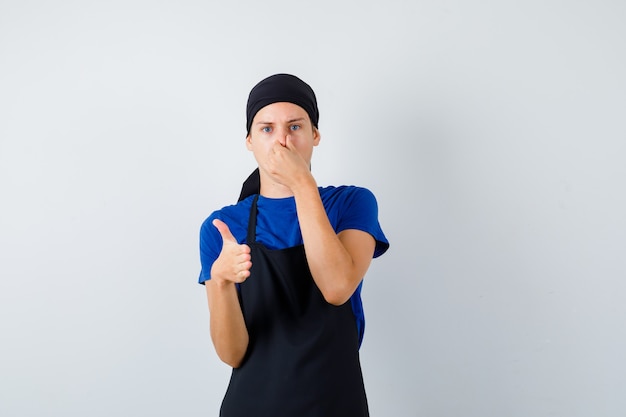 Retrato de cocinero adolescente masculino pellizcando la nariz y ofreciendo apretón de manos en camiseta, delantal y mirando disgustado vista frontal