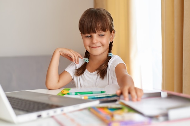 Retrato de colegial sonriente feliz, vestido con camiseta blanca sentado en la mesa contra la ventana con cortinas delante de la computadora portátil, con expresión positiva, haciendo los deberes.