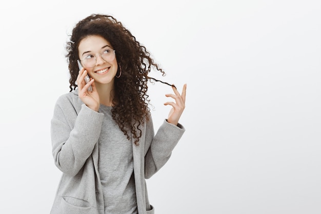 Retrato de una compañera de trabajo caucásica de buen aspecto coqueto en abrigo gris y gafas, mirando a un lado con una amplia sonrisa mientras habla por teléfono inteligente y juega con curl