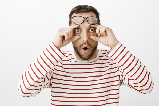 Retrato de un compañero de trabajo divertido sorprendido sorprendido, sosteniendo gafas en la frente y tirando de los párpados, mirando con los ojos abiertos algo extraño e increíble