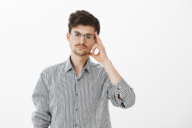 Retrato de un compañero de trabajo masculino serio y concentrado con gafas redondas, mirando hacia abajo y sosteniendo la sien con el dedo índice, concentrándose mientras piensa, inventando un plan sobre cómo evitar una situación incómoda