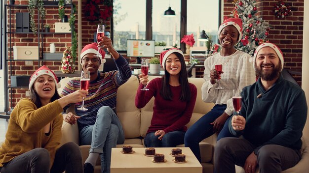 Retrato de compañeros de trabajo festivos tintineando vasos con alcohol para celebrar las fiestas navideñas con brindis o vítores. Gente celebrando el evento de fiesta de invierno con bebida de vino en una oficina decorada.