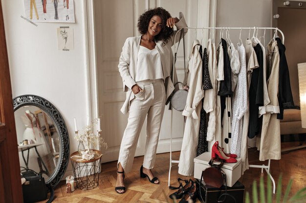 Retrato completo de una elegante mujer rizada de piel oscura con pantalones blancos, chaqueta y top posando en el vestidor La joven se apoya en la percha