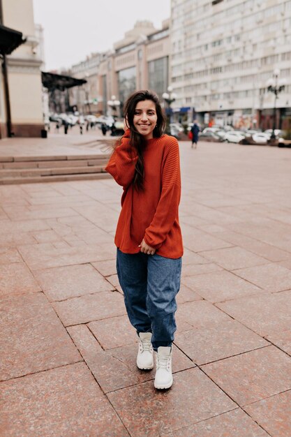 El retrato completo de una encantadora chica bonita con el pelo largo y oscuro y una sonrisa maravillosa lleva un suéter de punto naranja y jeans caminando por la calle en un día cálido