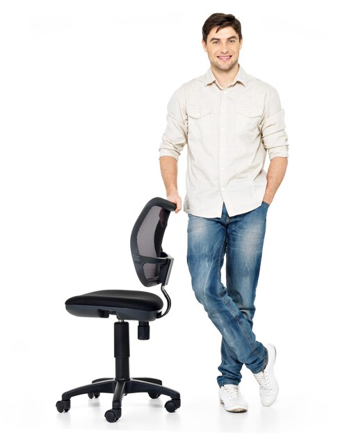 El retrato completo del hombre feliz sonriente se coloca cerca de la silla aislada en blanco.