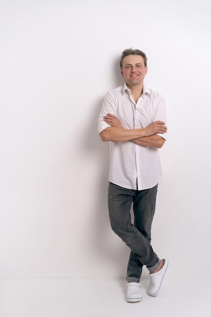 Retrato completo de un joven apuesto con camisa blanca y anteojos sonriendo con una expresión positiva feliz y segura con los brazos cruzados en una sesión de estudio de fondo blanco