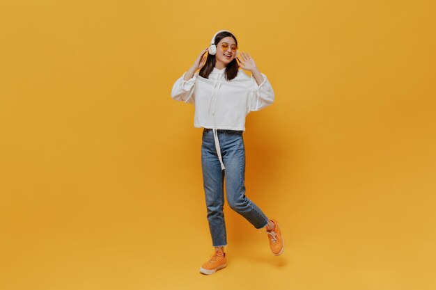 Retrato completo de mujer morena en pantalones de mezclilla con capucha blanca y gafas de sol naranjas cantando y escuchando música en auriculares sobre fondo naranja