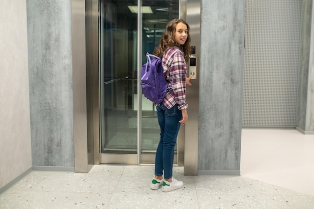 Retrato completo de un niño tranquilo y sonriente parado frente a la puerta del ascensor y presionando el botón de llamada