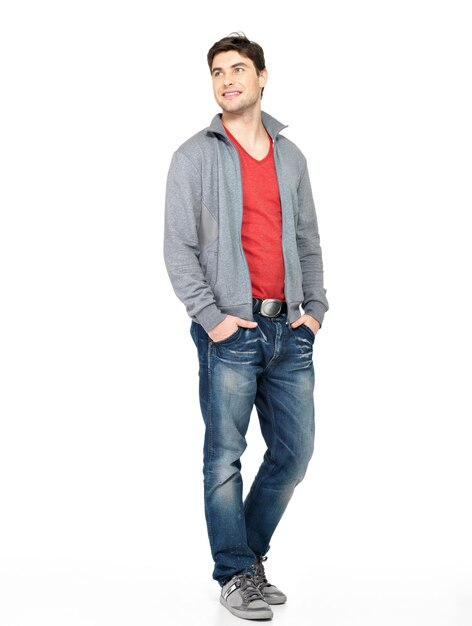 Retrato completo de sonriente feliz guapo en chaqueta gris, jeans. Hermoso chico de pie aislado en blanco mirando lejos