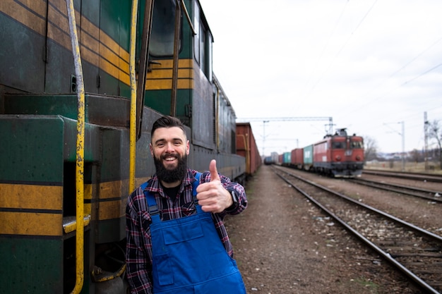Retrato del conductor del tren del motor de pie junto a la locomotora en la estación de tren y sosteniendo Thumbs up