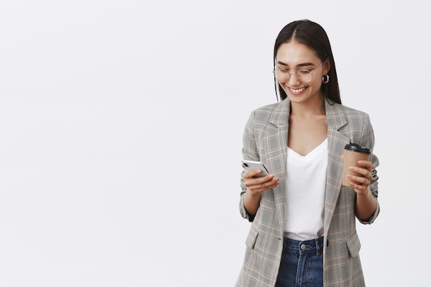 Retrato de confiada empresaria ocupada en gafas y chaqueta, sosteniendo una taza de café y un teléfono inteligente, usando gadget