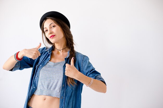 Retrato confiado joven de la mujer que lleva el sombrero que muestra los pulgares para arriba