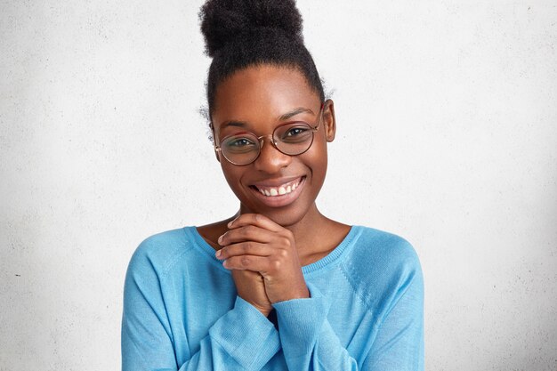 Retrato de contenido hermosa modelo femenina afroamericana sonríe con alegría y mantiene las manos juntas, contento de recibir felicitaciones