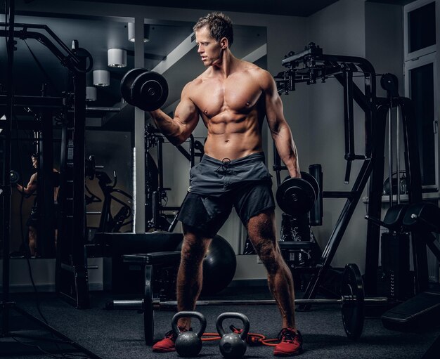 Retrato corporal completo de un hombre atlético sin camisa haciendo ejercicios de bíceps con pesas en un club de gimnasia.