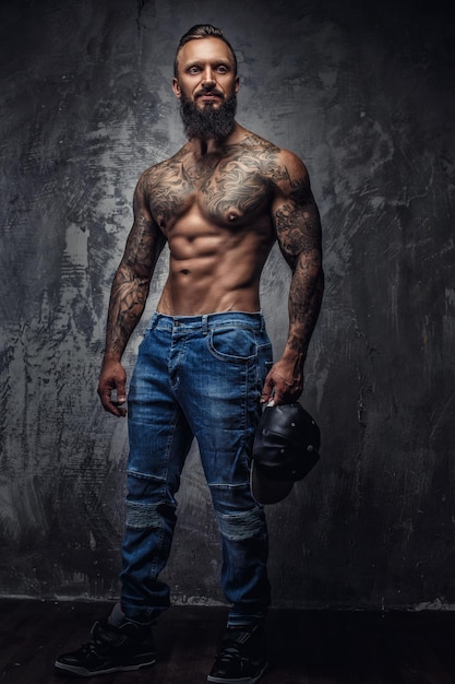 Retrato corporal completo de un hombre barbudo musculoso tatuado en jeans azules sobre una pared gris.