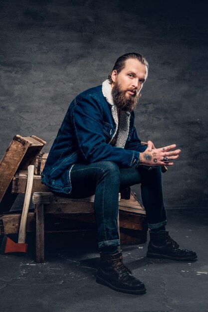 Retrato corporal completo de un hombre hipster barbudo que lleva una chaqueta de mezclilla se sienta en una caja de madera.