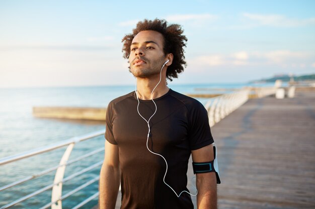 Retrato de corredor de hombre musculoso de piel oscura en forma con peinado tupido que parece concentrado en ropa deportiva negra con auriculares blancos.