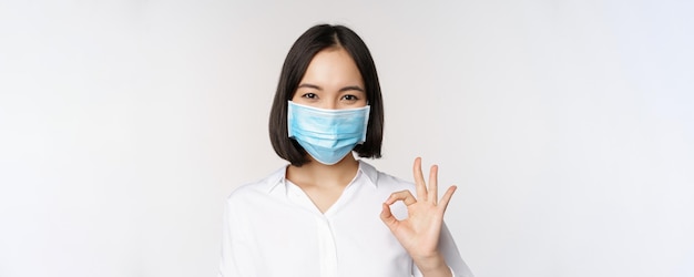 Retrato de covid y concepto de salud de una mujer asiática con mascarilla médica y mostrando un signo de aprobación sta