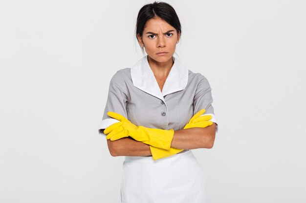 Retrato de criada atractiva gruñón en uniforme y guantes de goma amarilla de pie con las manos cruzadas