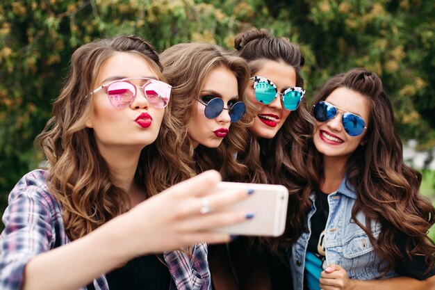 Retrato de cuatro novias con ropa informal de moda, peinados y maquillaje posando mientras hacen un autorretrato a través de un teléfono celular sentados afuera en el parque el día.