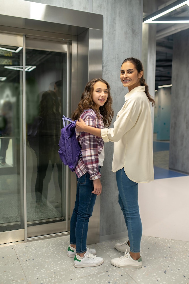 Foto gratuito retrato de cuerpo entero de un adolescente alegre y una señora sonriente de pie junto a las puertas cerradas del ascensor