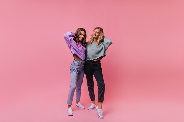 Retrato de cuerpo entero de una agradable chica morena en jeans pasar tiempo con su hermana. Señoras despreocupadas en traje casual aislado en pastel.