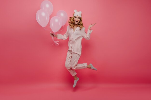 Retrato de cuerpo entero de una agradable cumpleañera en calcetines saltando sobre la pared rosa. Linda mujer joven en pijama y antifaz divirtiéndose con globos de helio.