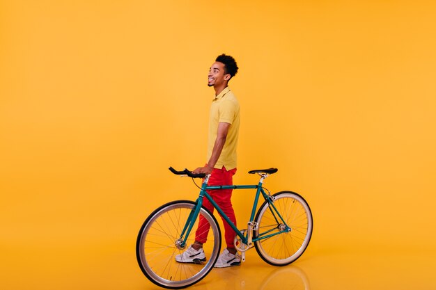 Retrato de cuerpo entero de alegre hombre africano posando con bicicleta. Foto de chico negro de moda en calzado deportivo de pie.