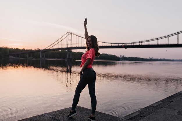 Retrato de cuerpo entero de una chica deportiva disfrutando de las vistas al río en la noche. Retrato al aire libre de una impresionante dama sonriente haciendo fitness en la naturaleza.