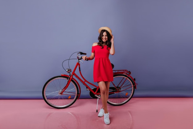 Retrato de cuerpo entero de una chica elegante con vestido rojo de verano de pie en el estudio sobre fondo púrpura Mujer morena despreocupada con sombrero de paja posando con su bicicleta