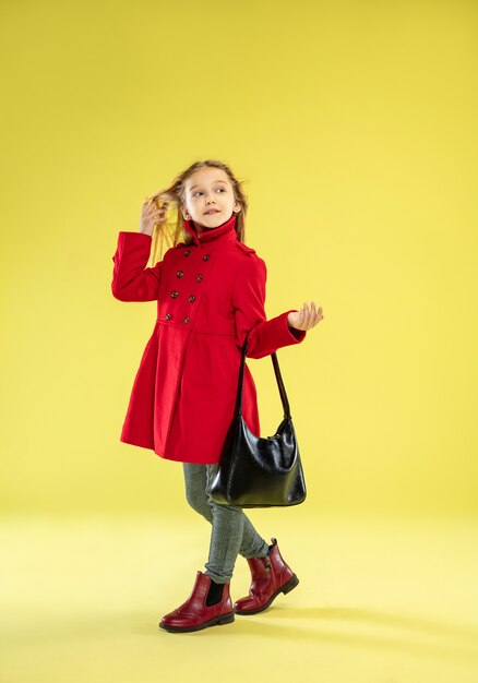 Un retrato de cuerpo entero de una chica de moda brillante en un impermeable rojo con bolso negro posando en la pared amarilla del estudio