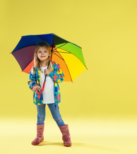 Un retrato de cuerpo entero de una chica de moda brillante en un impermeable sosteniendo un paraguas de colores del arco iris sobre fondo amarillo de estudio.