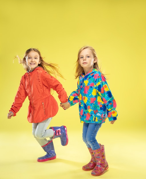 Un retrato de cuerpo entero de chicas de moda brillantes en un impermeable tomados de la mano, corriendo y divirtiéndose en la pared amarilla del estudio