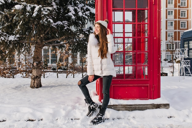 Retrato de cuerpo entero de una dama entusiasta con peinado largo posando junto a la caja de llamada roja en invierno. Foto exterior de mujer bonita caucásica con sombrero blanco disfrutando de las vacaciones de diciembre en Inglaterra.