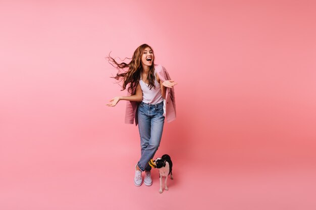 Retrato de cuerpo entero de una dama morena en jeans posando con perro. Retrato de interior de hermosa modelo femenina de pie junto a bulldog francés.
