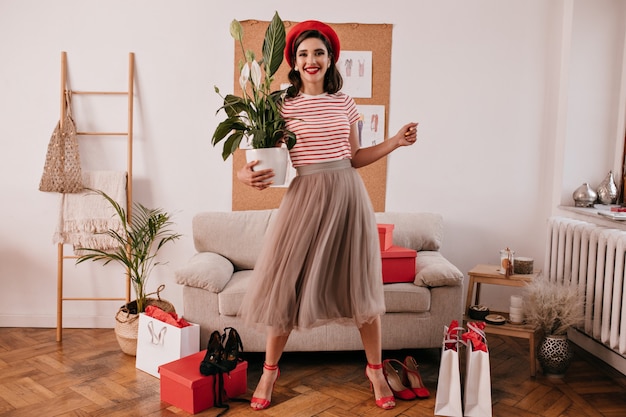 Retrato de cuerpo entero de dama en vestido posando con planta. Joven mujer moderna en ropa elegante mira a cámara y sonríe.