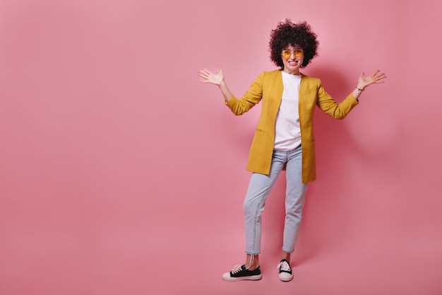 Retrato de cuerpo entero de divertido modelo de mujer joven con rizos cortos vistiendo chaqueta amarilla y jeans sonriendo al frente