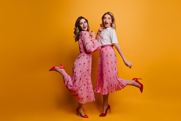 Retrato de cuerpo entero de dos amigas lleva zapatos rojos de tacón alto. Foto interior de damas entusiastas relajándose juntas.