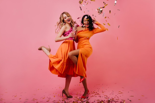Retrato de cuerpo entero de dos chicas bailando bajo confeti Foto de estudio de mujeres americanas disfrutando del evento