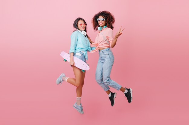 Retrato de cuerpo entero de dos mujeres deportivas saltando y sonriendo. Chica glamorosa patinadora en camisa azul divirtiéndose con una amiga africana en zapatos negros.