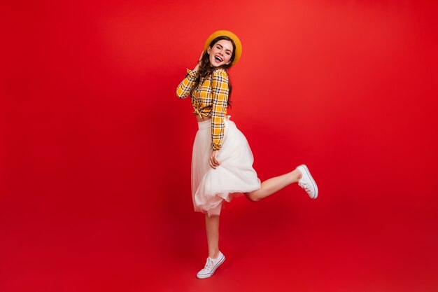 Retrato de cuerpo entero de una elegante dama positiva saltando sobre la pared roja. Mujer con camisa a cuadros y falda blanca está bailando de muy buen humor.